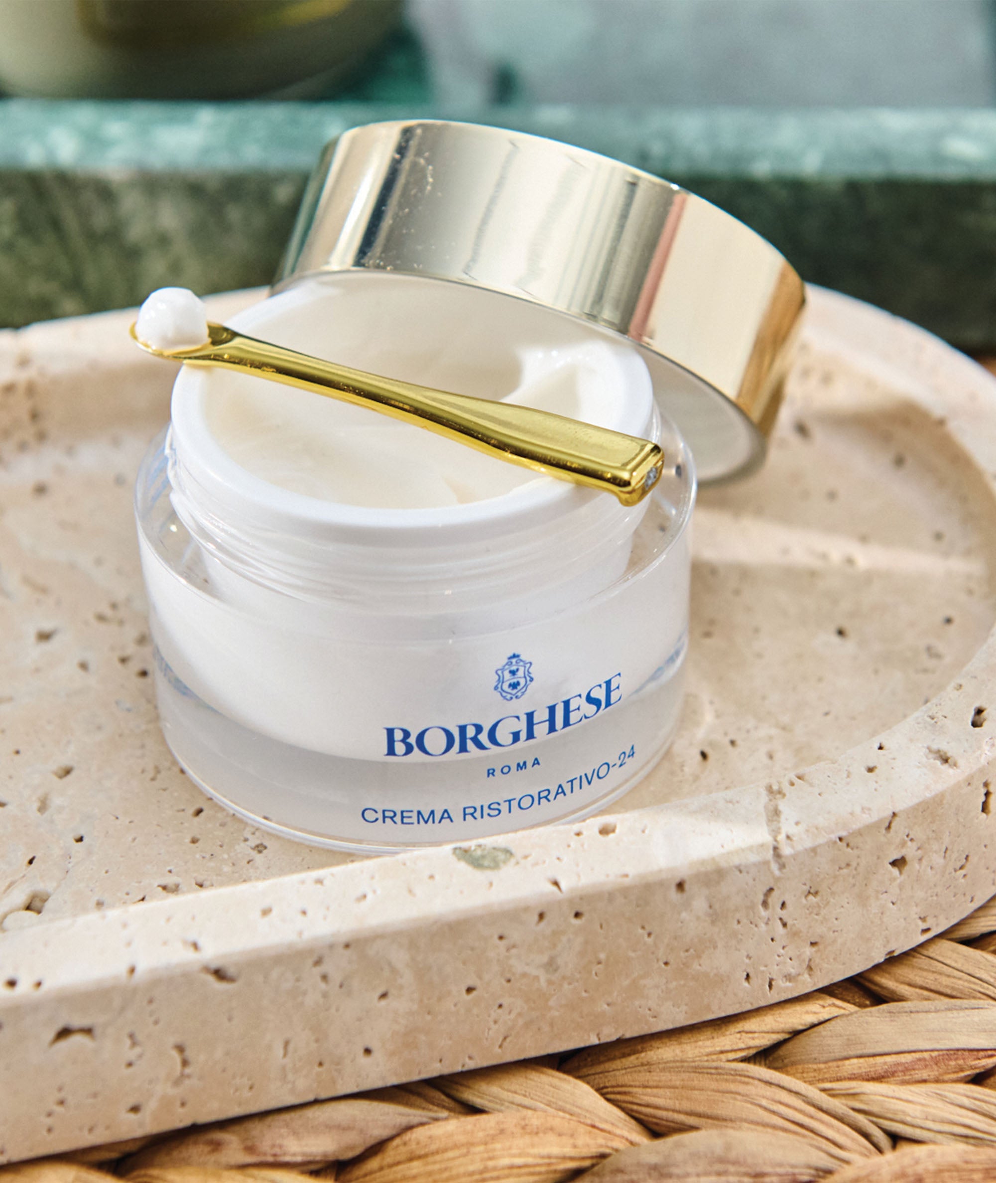 Lifestyle image of Crema Ristorativo-24 Moisturizer with a lid propped on a stone tray showing a spatula with pea sized amount 